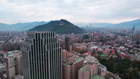 Panorama-Luftaufnahme-Von-Santiago,-Chile,-Mit-Dem-Centenario-Turm-Im-Vordergrund-Und-Dem-Hügel-San-Cristobal-Und-Den-Anden-Im-Wolkigen-Hintergrund