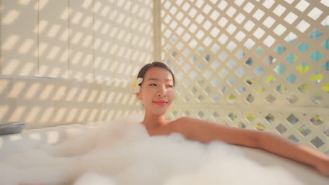 Beautiful-Asian-woman-wearing-a-frangipani-plumeria-flower-on-her-ear-enjoys-a-soothing-bubble-bath-in-a-tub