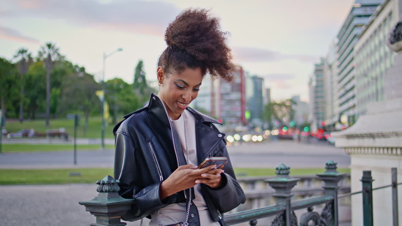 Cheerful woman messaging cellphone leaning on urban railings at evening closeup