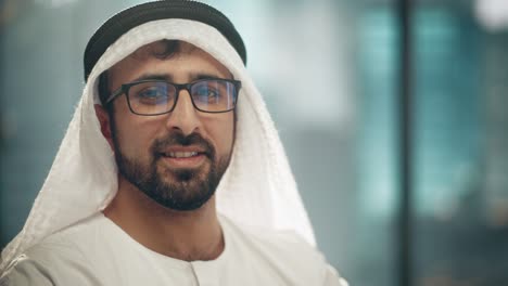 portrait of successful arab businessman in traditional outfit gently smiling, wearing glasses, white kandura and black agal keeping a ghutra in place. saudi, emirati, arab businessman concept.