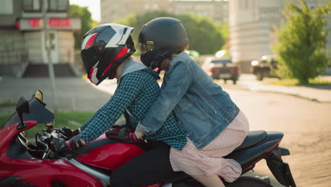 dos hermanas montan una bicicleta eléctrica roja con cascos mientras pasan por una ciudad urbana, el fondo presenta un borrón de edificios, coches estacionados y personas caminando