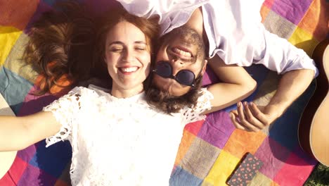 feliz y encantadora pareja joven acostada en una manta de colores en el parque mirando al cielo apuntando a las nubes romántico con gafas de sol de guitarra hermosa chica atractiva vista superior cámara giratoria en cámara lenta