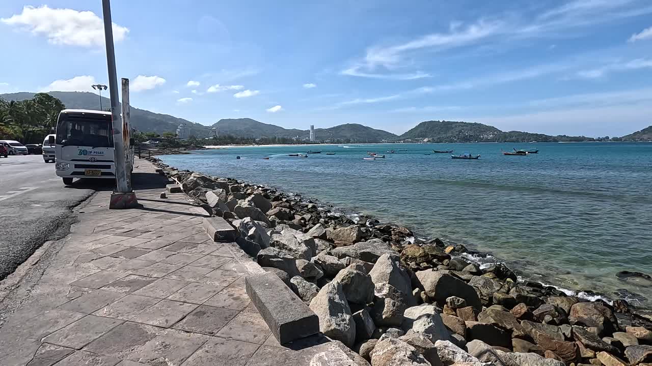 A tranquil coastal scene with clear skies, rocky shoreline, and parked vehicles in Phuket, Thailand. Captured in natural daylight
