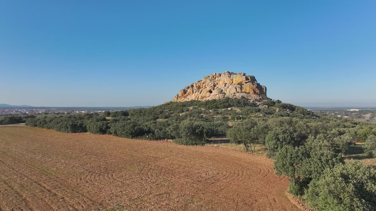 Drone ascending over plowed farmland with scattered holm oaks, flying frontal towards a striking rocky hill. The vivid colors of the formation frame a dramatic cave entrance in the landscape