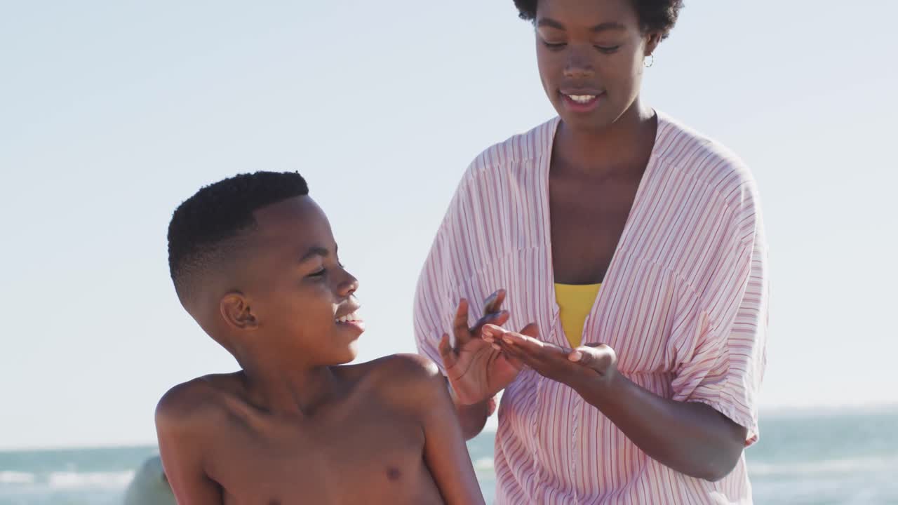 Video of happy african american mother putting sunscreen on her son on beach