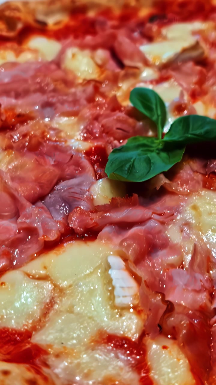 Detailní pohled na čerstvě upečenou pizzu s rozpuštěným sýrem, šunkou a čerstvými lístky bazalky.