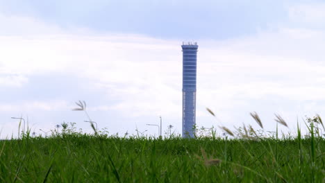 Air-Traffic-Controller-Tower-seen-from-a-distant-horizon-with-land-covered-with-grass-in-the-foreground-during-a-windy-and-cloudy-day-in-Bangkok,-Thailand