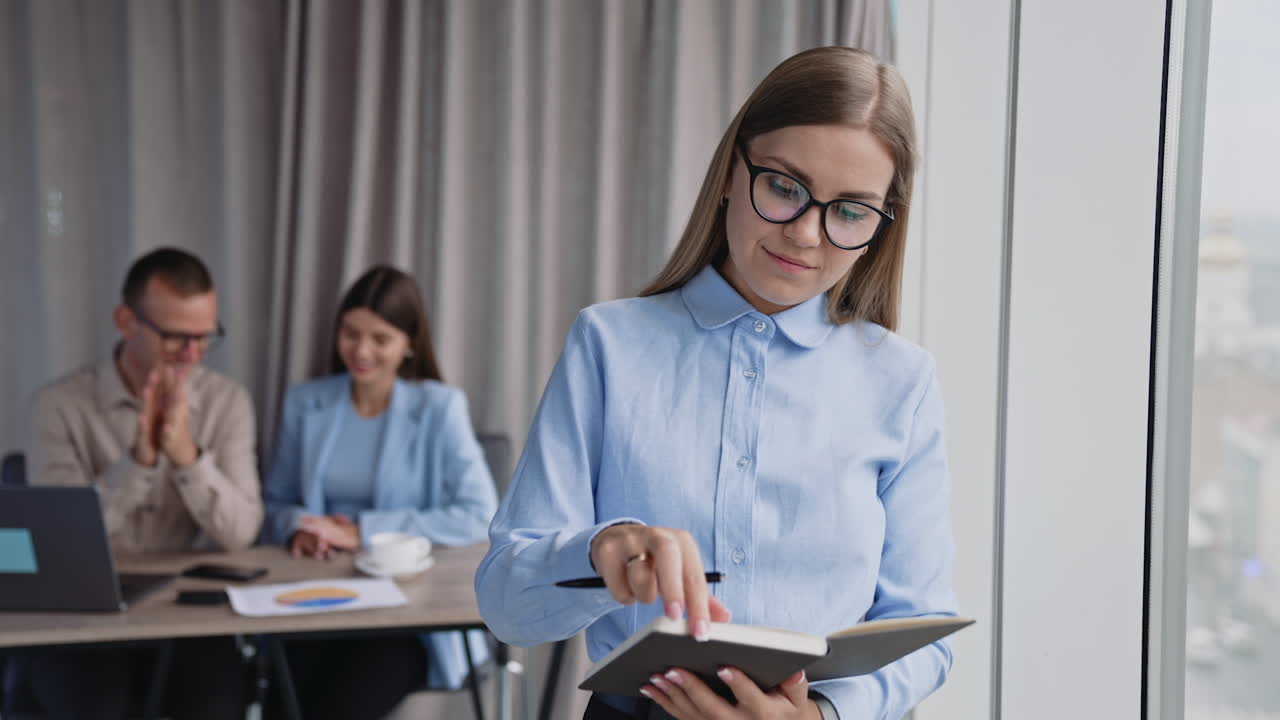 Blonde lady in glasses taking notes into her paper notebook standing at the window. Couple of colleagues sitting at desk behind in blur.