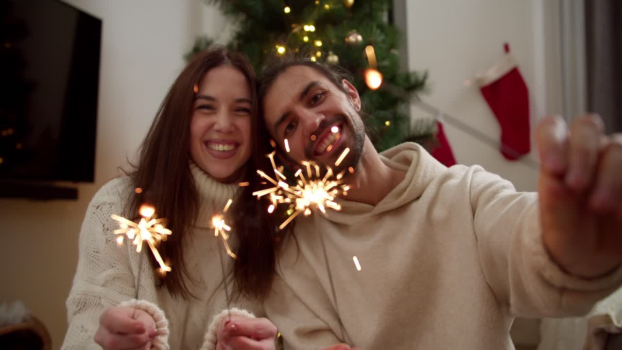 Portrait: a happy couple, a guy and a brunette girl in white sweaters, looks at the camera and smiles and holds sparklers in their hands that glow brightly and burn in the New Year's atmosphere near the New Year's tree in a cozy home in winter