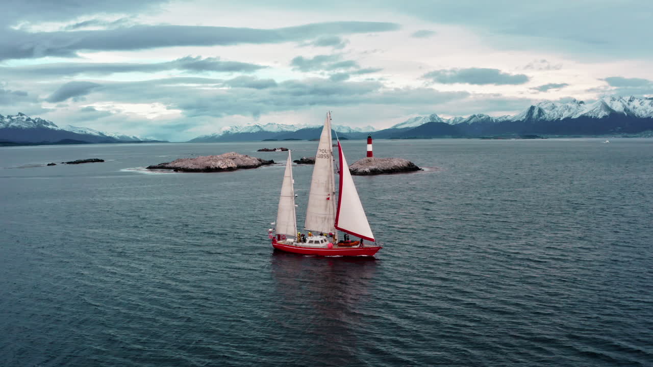Red sailboat sails solo on wide Patagonian channel with gray sky and mountain background, aerial tracking right