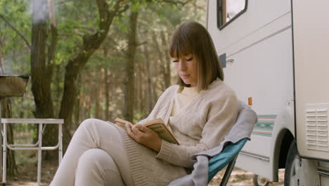 mulher concentrada lendo um livro enquanto está sentada em uma cadeira perto de um trailer na floresta