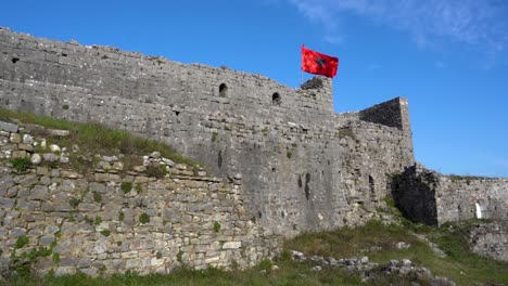 Stone-surrounding-walls-of-Rozafa-castle-and-Albanian-flag-waving-in-blue-sky-background