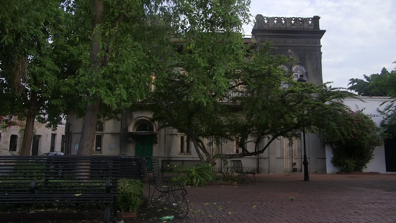 Patio histórico en Santo Domingo, República Dominicana, con arquitectura de la era colonial, pavimento de ladrillo, bancos y exuberantes árboles tropicales en un tranquilo entorno urbano.