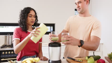 making smoothies, young multiracial couple pouring green smoothie into glasses in kitchen, at home