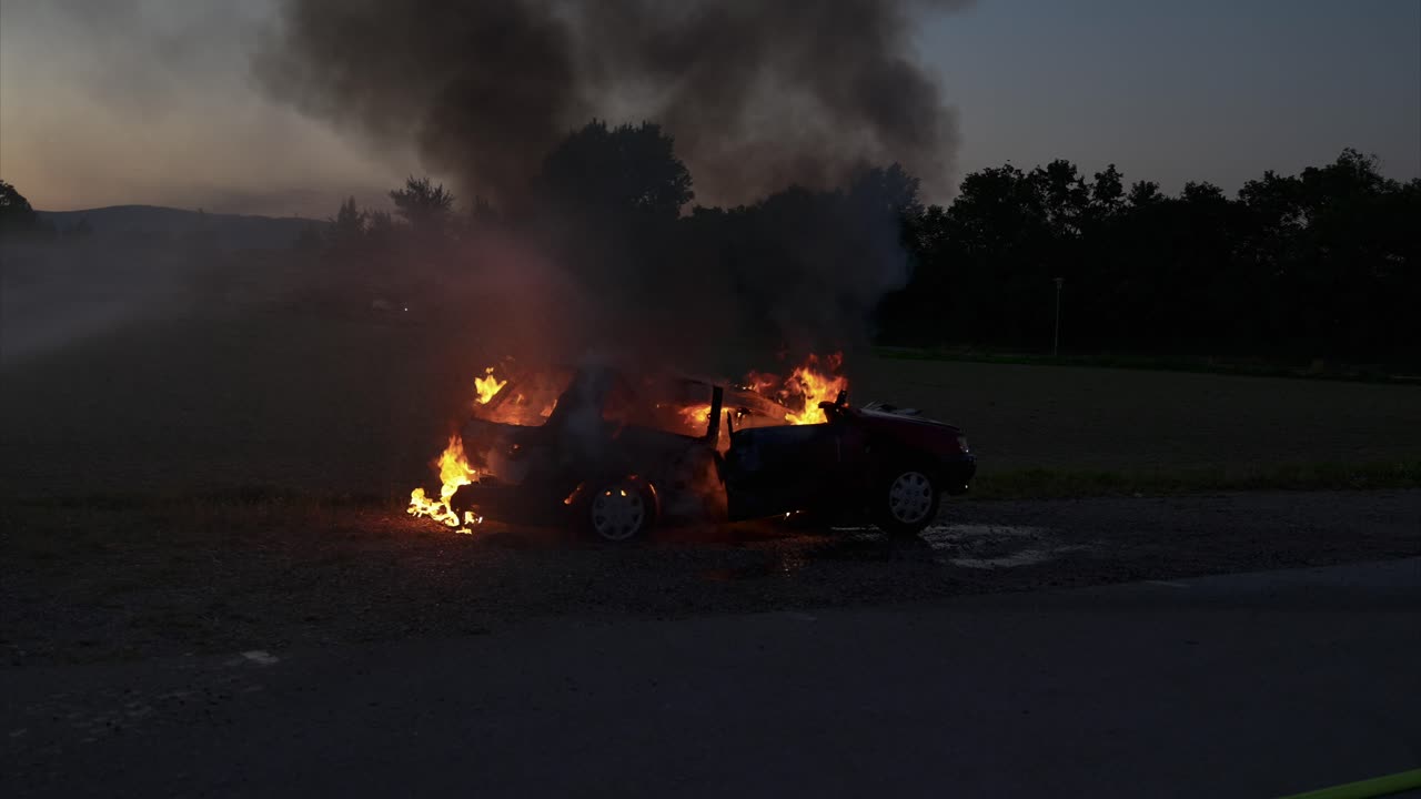 Burning car in the evening hours is extinguished with water from the left - the fire is already almost gone