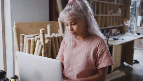 Young-Female-Business-Owner-In-Workshop-For-Sustainable-Bicycles-Doing-Accounts-On-Laptop