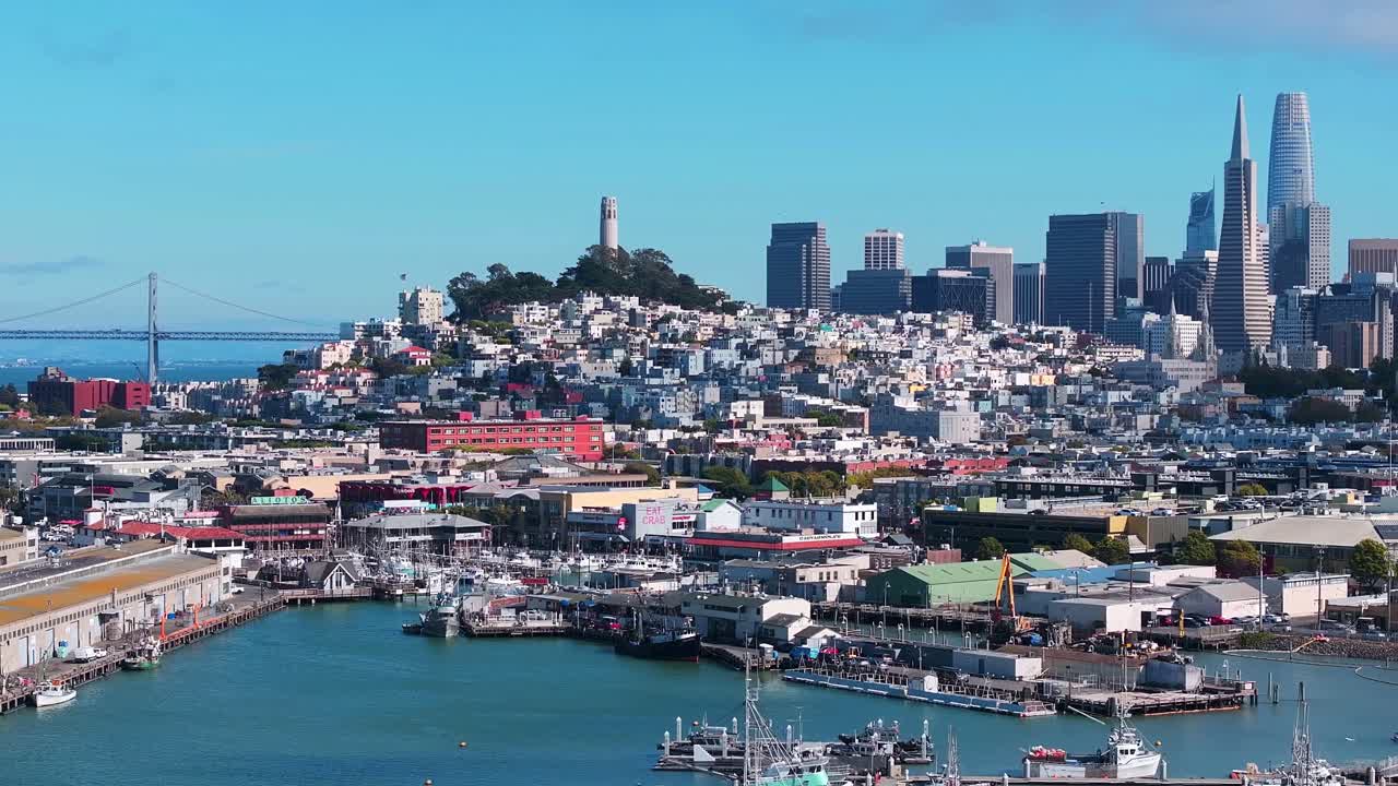 Wide daytime view of San Francisco, California, with Fisherman's Wharf marina in the foreground, Coit Tower on Telegraph Hill, downtown skyline, and the Bay Bridge under a clear blue sky