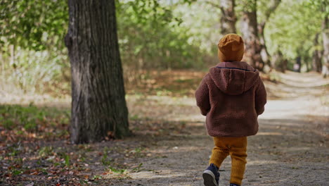 lovely toddler wearing yellow cap, suit and warm jacket walks in the park. baby boy runs by the path pointing up with a finger.