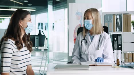 doctora caucásica con mascarilla médica sentada en el escritorio y explicando a una paciente el tratamiento del coronavirus en consulta médica