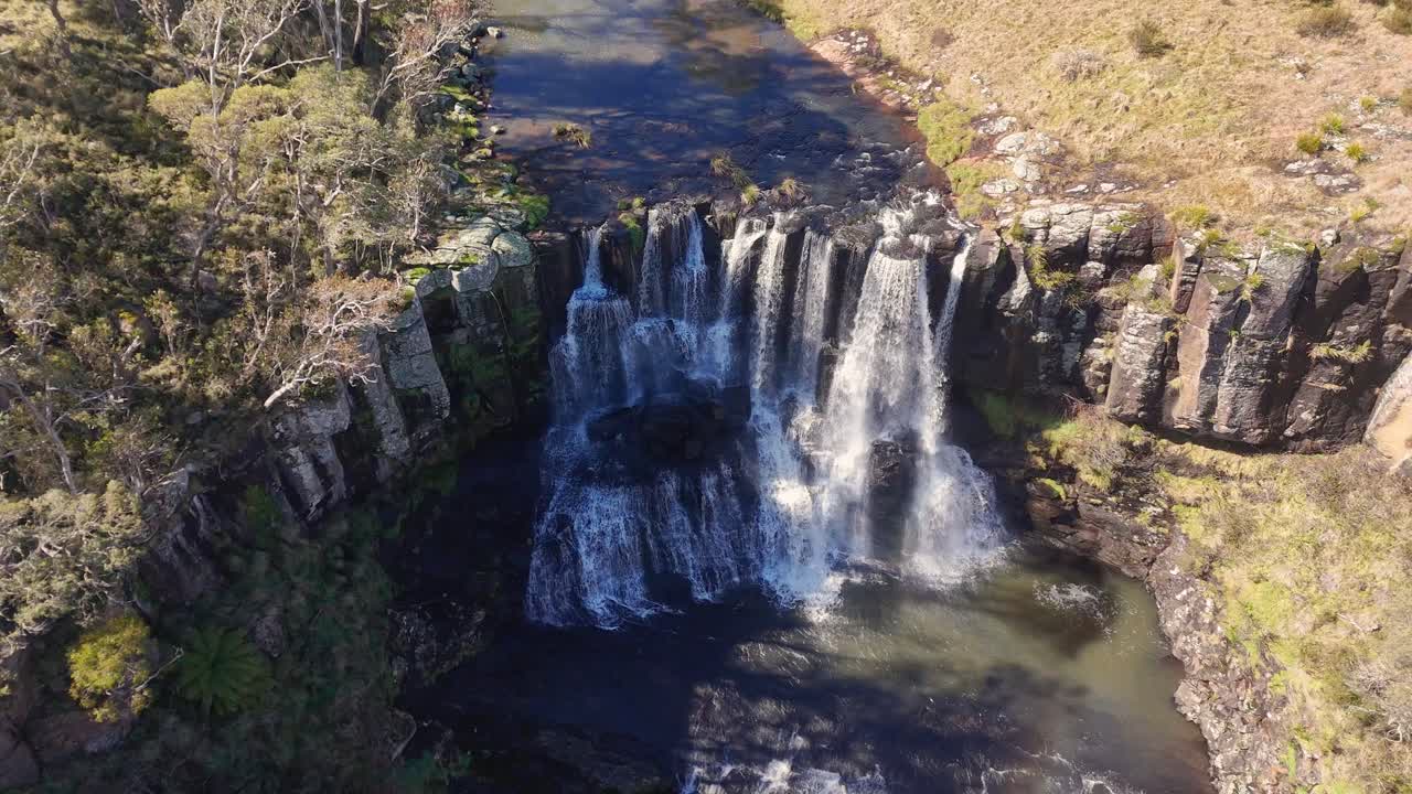 Drone footage captures Ebor Falls in New South Wales, Australia, with water cascading over cliffs, surrounded by forest and bathed in natural daylight