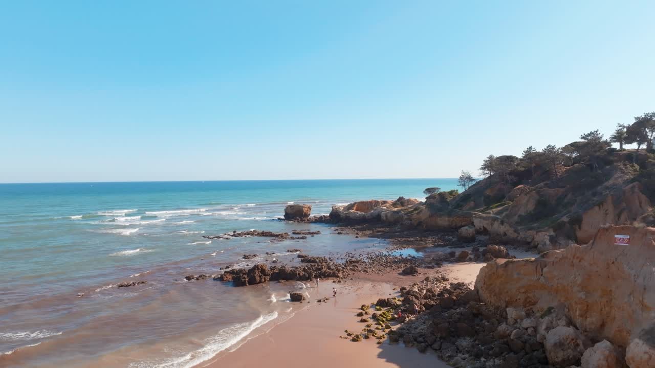 Pessoas passeiam em praia vazia brincando na água perto da costa rochosa, Praia da Santa Eulália, Albufeira, Algarve