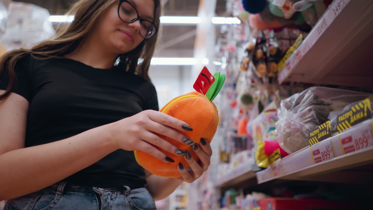 primer plano de una mujer con gafas seleccionando un bolso naranja y verde brillante de un estante de la tienda, la compradora navega por accesorios elegantes en un entorno minorista