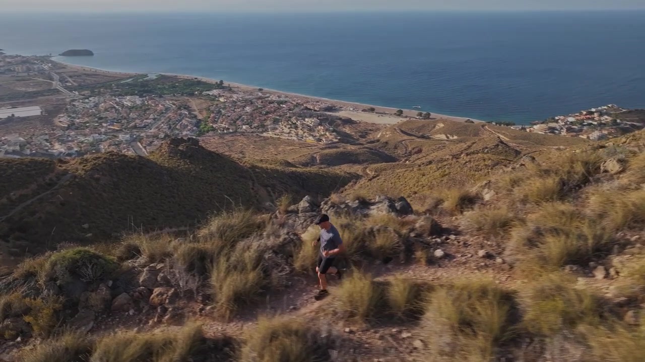 Ripresa aerea dall'alto che segue un uomo che corre in discesa lungo uno stretto sentiero di crinale montano, con vista su una pittoresca città costiera e sul mare.