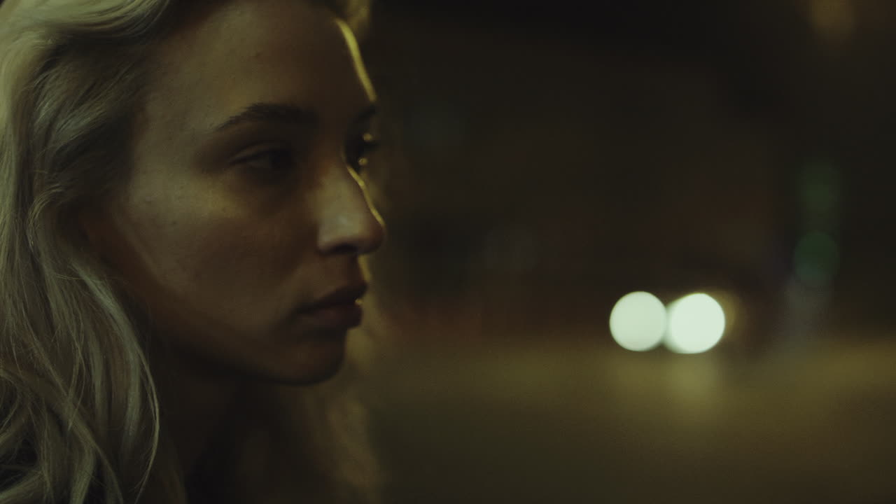 Blonde woman city night face looking at cars passing in street lights.