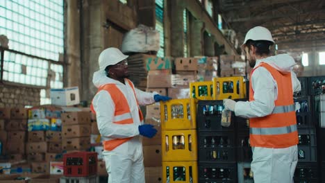 A-man-with-Black-skin-in-a-white-protective-uniform-together-with-his-colleague-a-brunette-man-with-a-beard-in-an-orange-vest-puts-glass-bottles-in-special-containers-and-communicates-while-they-work-at-a-waste-recycling-plant