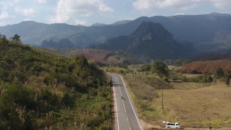 Vista-Panorámica-Aérea-De-Un-Motociclista-Cruzando-Una-Carretera-Solitaria-Entre-Montañas-En-El-Sudeste-Asiático