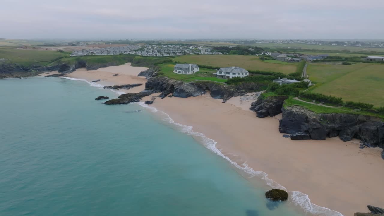 Pristine Cornish beach with clear blue water and houses on dark jagged sea cliffs next to caravan park. Mother Ivey's Bay, Cornwall, UK.
