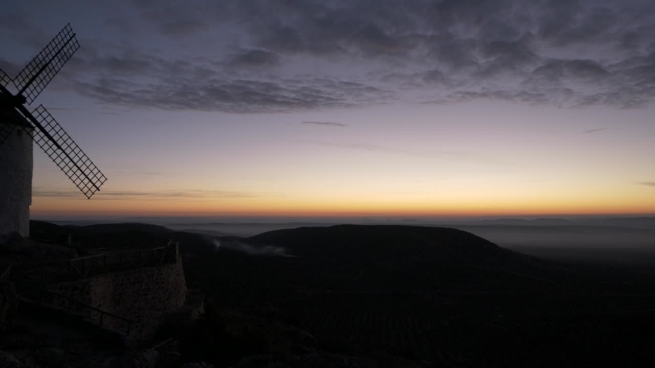 una panorámica hacia la derecha que comienza en un molino de viento desde la cima de una montaña en medio de una llanura, con niebla cerca del suelo en primer plano