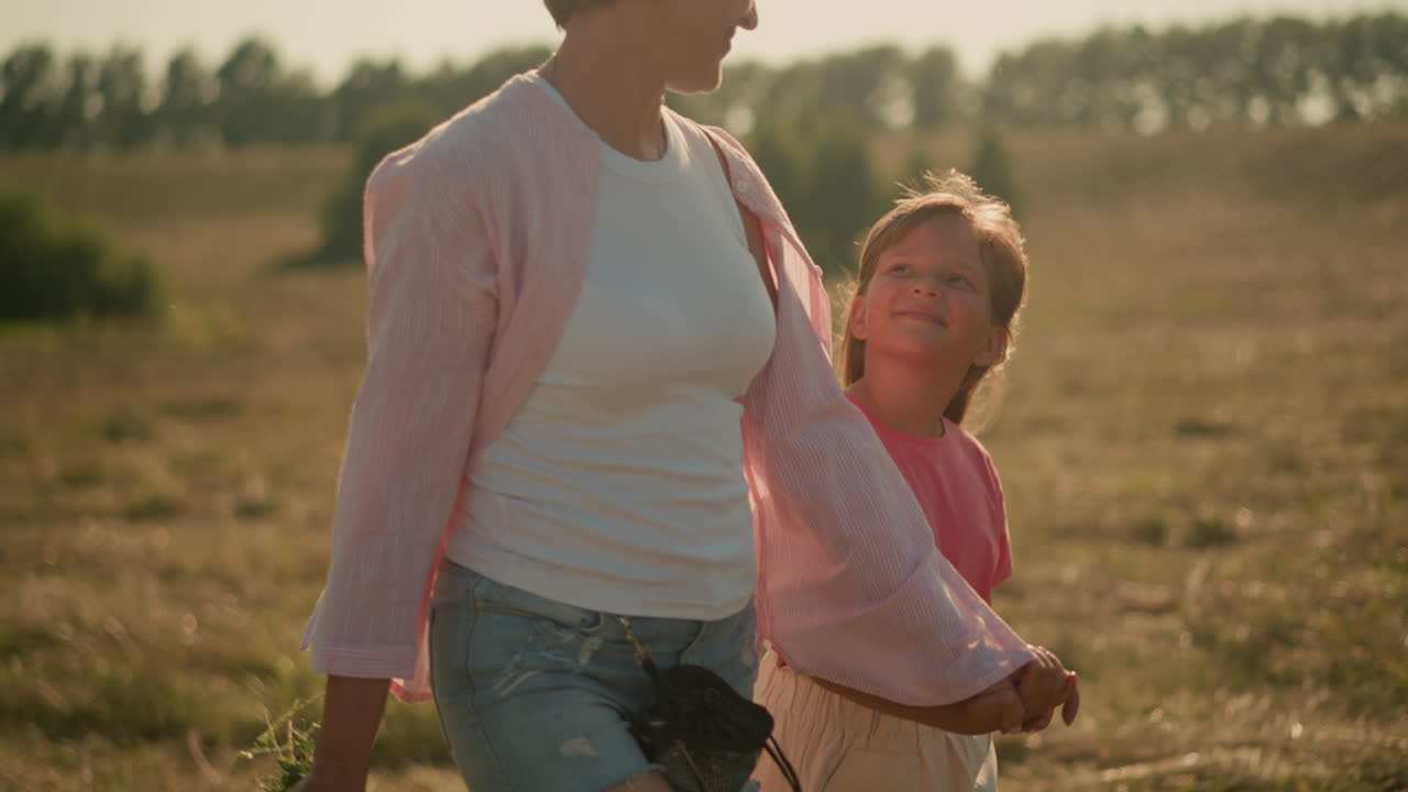 엄마와 딸이 손을 잡고 함께 걸어가며 빛 하늘 아래 즐거운 미소를 나누는 클로즈업, 엄마가 손에 꽃을 들고, 배경에는 흐릿한 자연 풍경