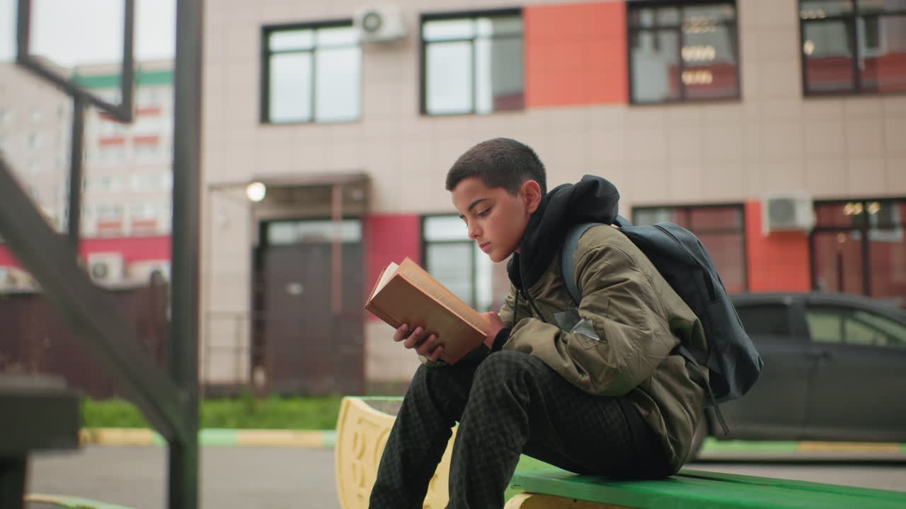 Student seated with backpack flipping through book while studying outdoors on green bench with blurred city building and glowing bulb above door in background