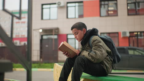student seated with backpack flipping through book while studying outdoors on green bench with blurred city building and glowing bulb above door in background