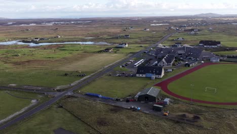 Wide-angle-drone-shot-of-Linaclate-in-Benbecula,-featuring-the-Dark-Island-Hotel,-the-local-UHI-college-campus,-the-nearby-football-pitch-and-running-track,-as-well-as-the-distant-moor-and-peatland