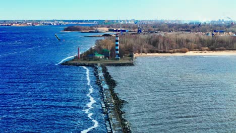 A tall striped lighthouse stands boldly on a weathered jetty, surrounded by vibrant blue waves and a windswept empty shoreline under crisp daylight.