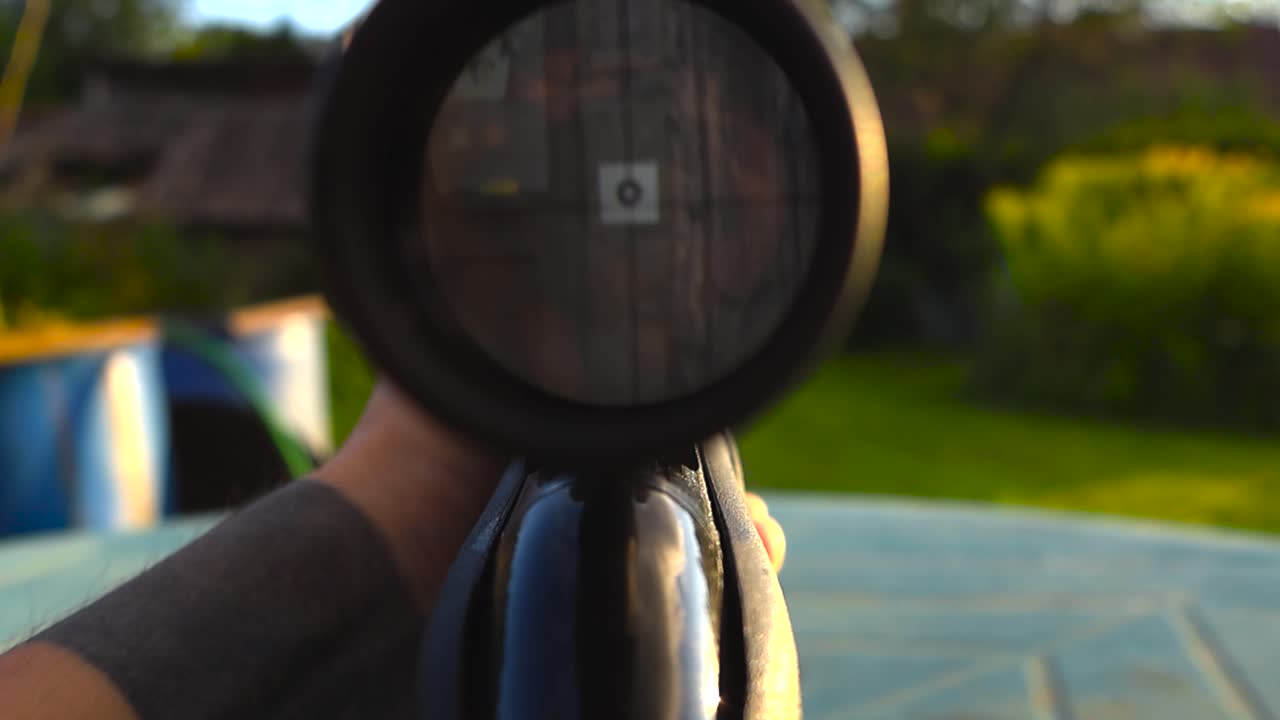 Point of view video of of a small shooting range mark viewed through a rifle weapon black colored scope or optics during a summer sunny day. Tattooed man white hand holding the weapon, target practice