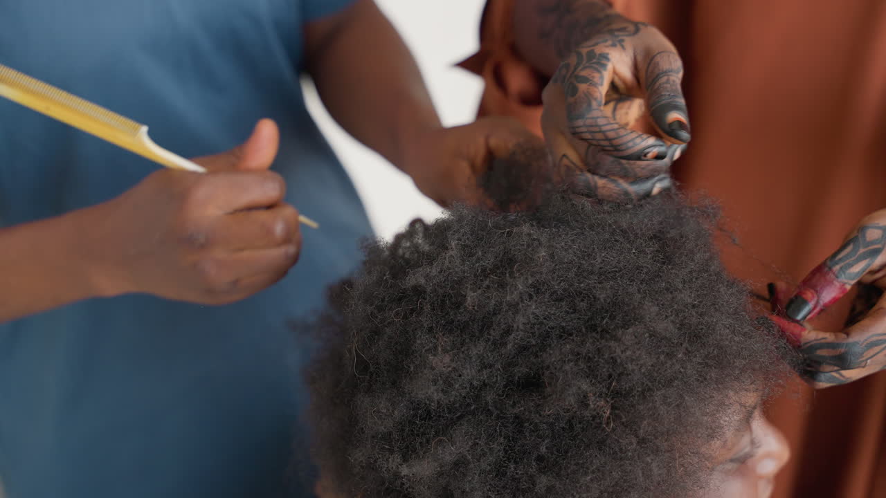Stilista acconcia delicatamente ricci africani. Primo piano della cura dei capelli con tatuaggi e smalto per unghie. Ambiente da studio interno che mostra un bambino con tatuaggi artistici e capelli afro accuratamente acconciati.