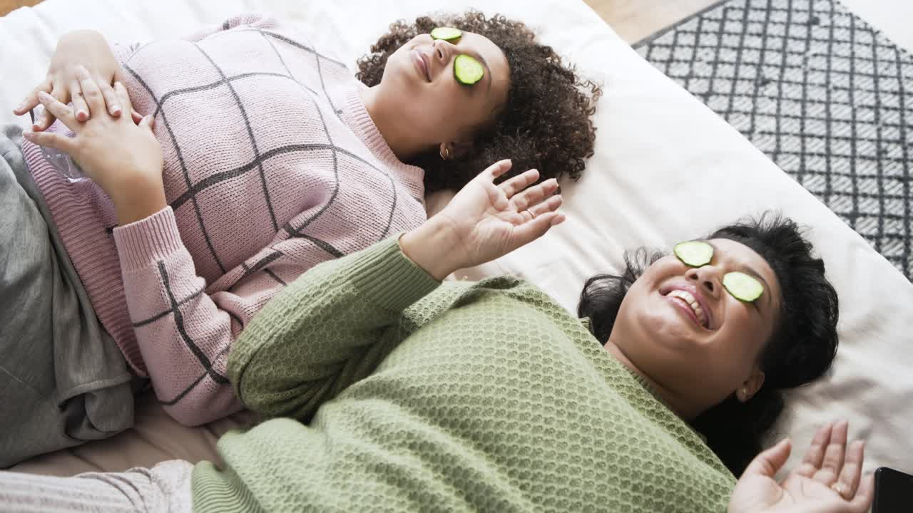 Grabbing cucumber slices African American female friends placing slices over eyes on bed relaxing