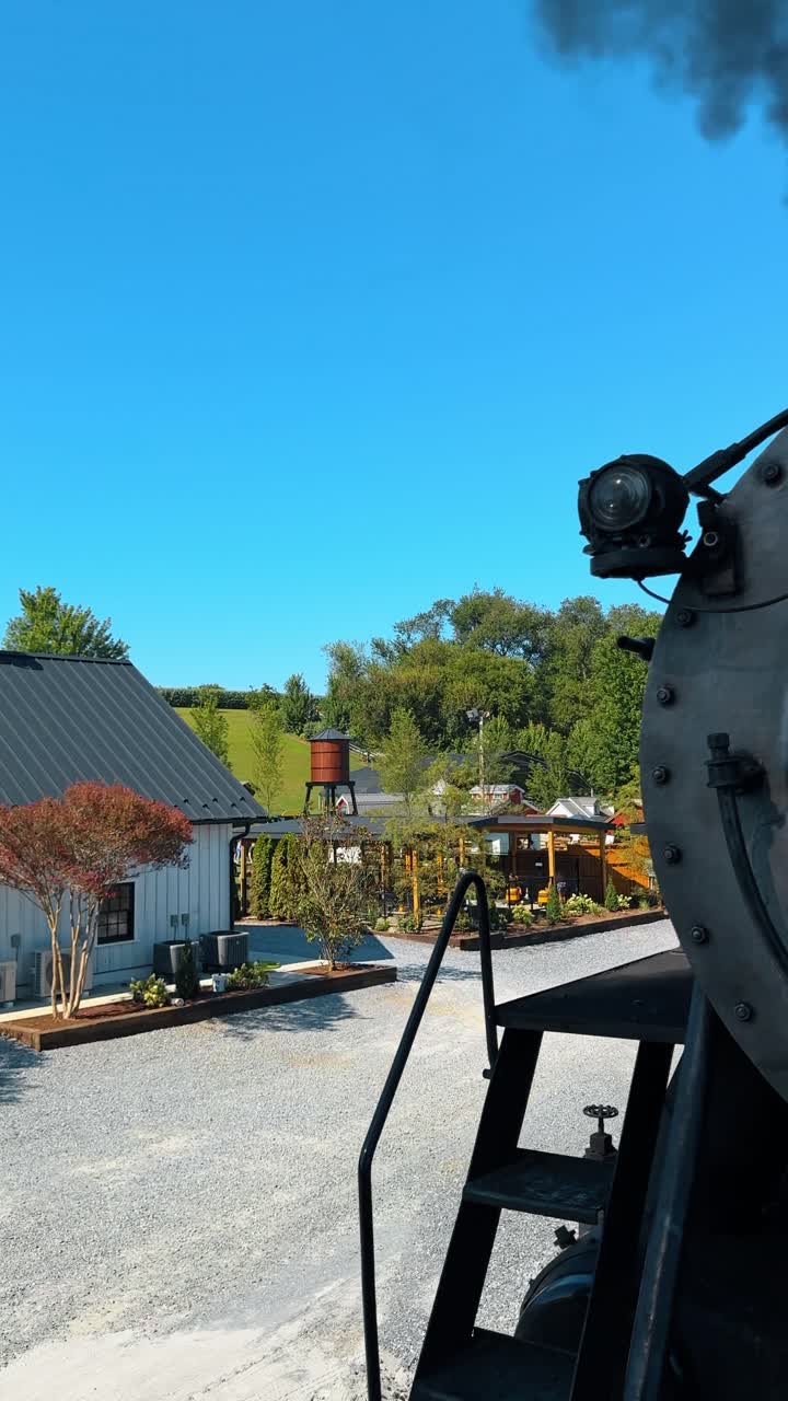 Udsigt fra damplokomotiv over jernbanemuseumsarealet. Perspektiv fra et veteran-damplokomotiv med udsigt mod et vandtårn i træ og hvide bygninger under en klar himmel.