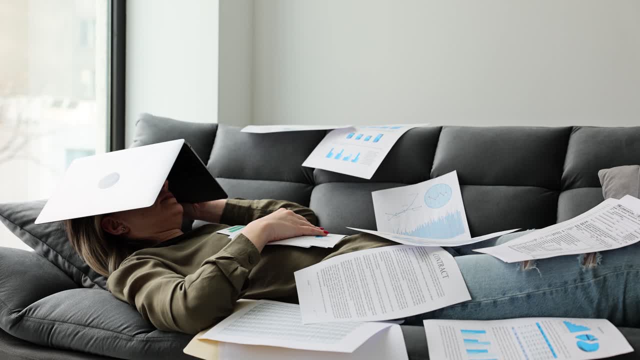 A stressed woman lies on a couch covered in work documents with a laptop on her head