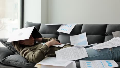 A stressed woman lies on a couch covered in work documents with a laptop on her head