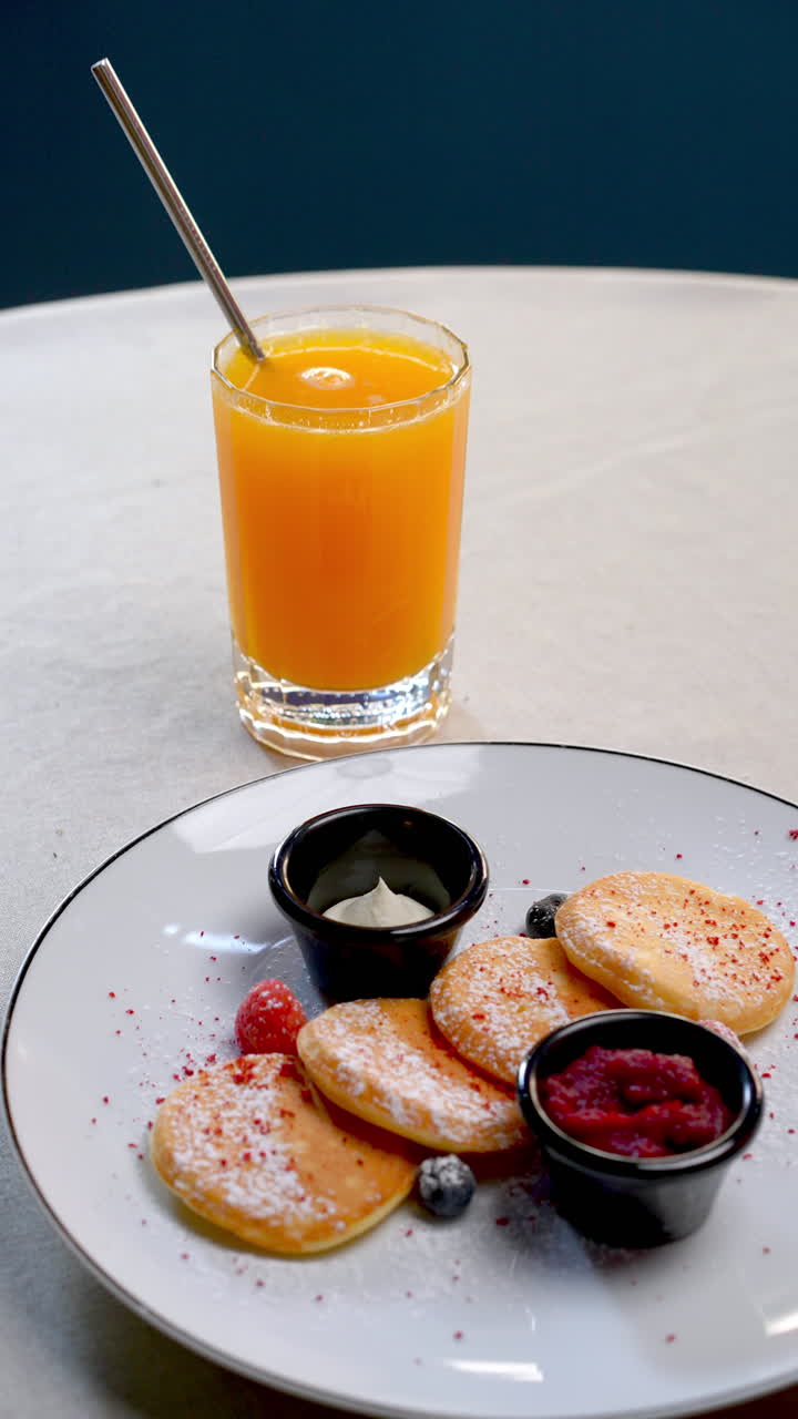 Small pancakes, covered with powdered sugar, served with fresh berries, jam, cream and a glass of orange juice, composing a delicious breakfast