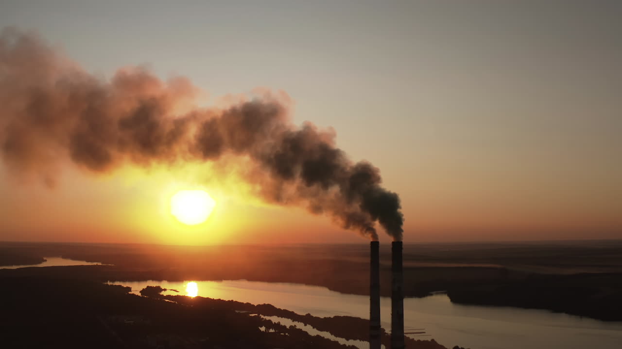 Factory pollutes environment at sunset. Toxic enterprise chimneys tubing against the evening sky background release black smoke. Smoking pipes of harmful manufacture in nature.
