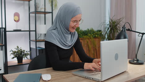 uma jovem de hijab e óculos está trabalhando em seu laptop em um escritório em casa.