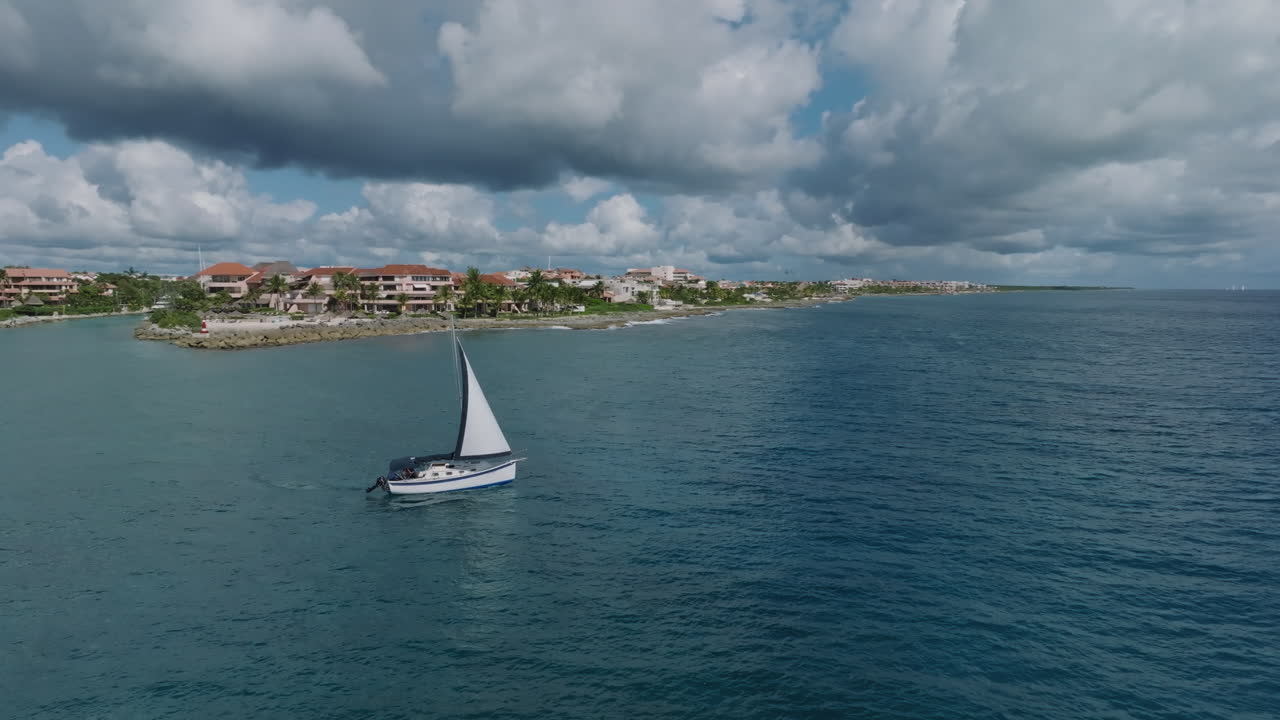 una impresionante toma aérea de rotación de un yate de vela que sale de la costa, revelando un impresionante edificio de apartamentos de vacaciones con una pintoresca orilla del mar en primer plano en puerto avenuras, méxico