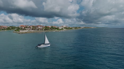 An-Stunning-Rotation-Aerial-Shot-of-a-Sailing-Yacht-leaving-the-Shoreline,-Revealing-a-Stunning-Holiday-Apartment-Building-with-a-Picturesque-Seashore-in-the-Foreground-in-Puerto-Avenuras,-Mexico