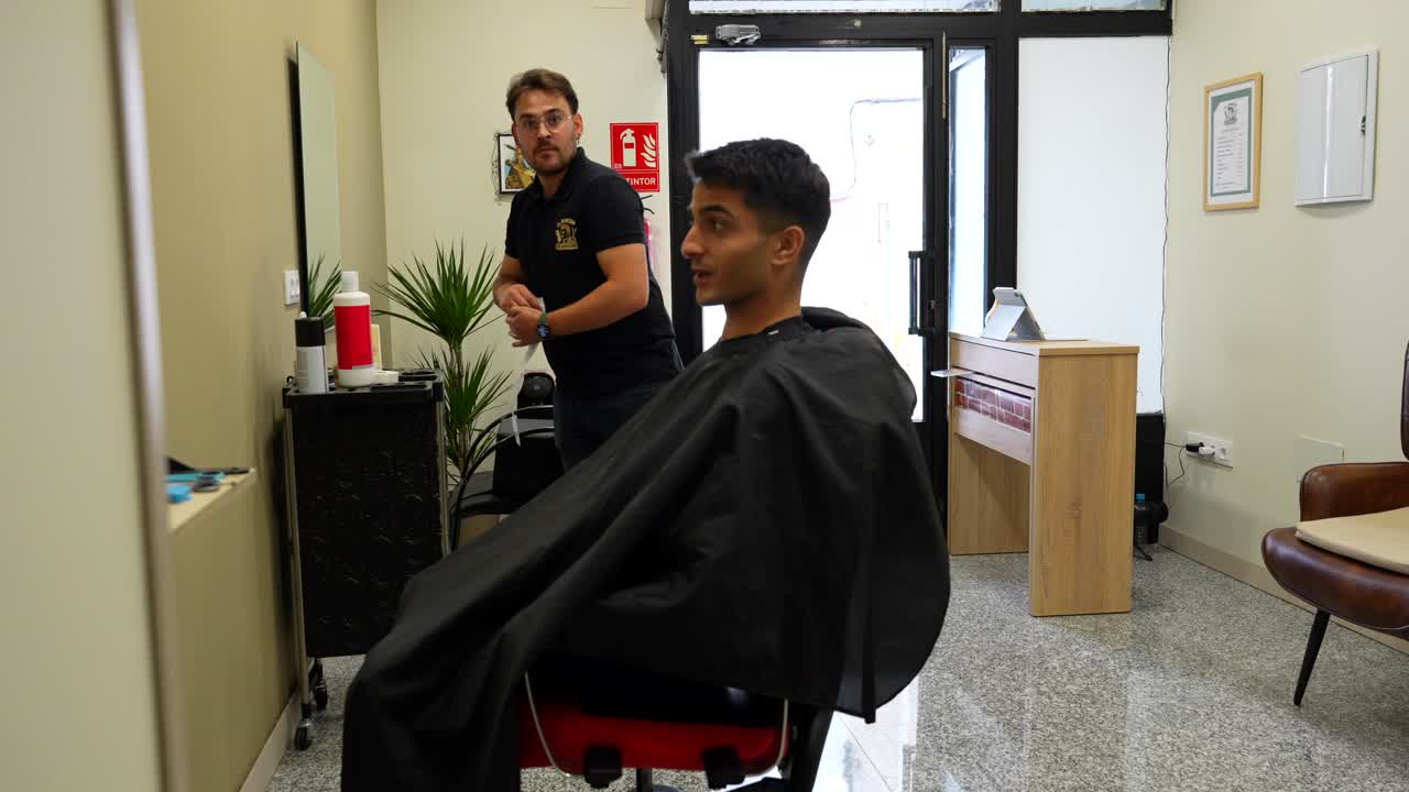 Kunde, der kigger sig i spejlet, mens barberen sætter en hvid halskrave på for at starte klipningen i en stilfuld barbersalon