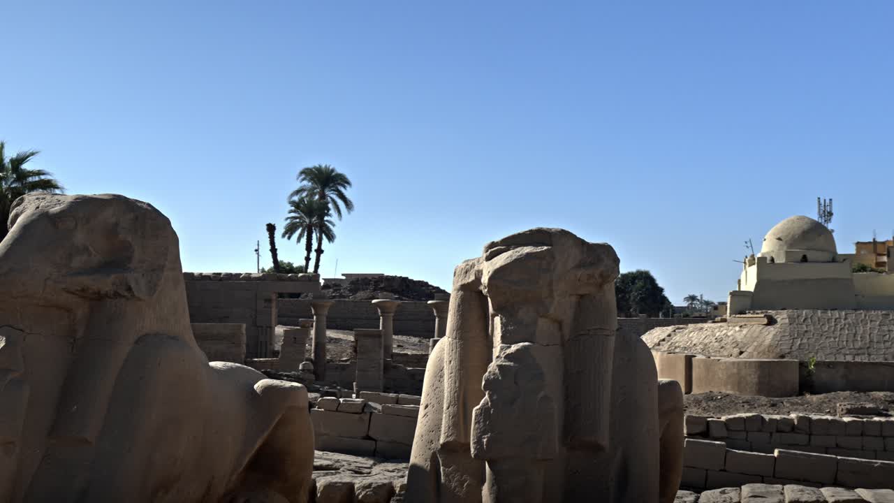 Situata nel Complesso del Tempio di Karnak a Luxor, Egitto, questa inquadratura ampia cattura la parte finale del Viale delle Sfingi (dromos) mentre si estende verso il canale moderno e il Nilo
